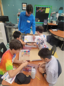 A man in a blue shirt assists four boys working on electronic projects at a classroom table.